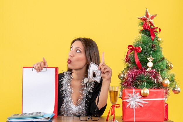 Estado de ánimo de año nuevo con una encantadora dama en traje sosteniendo una máscara y mostrando un documento pensando profundamente en la oficina