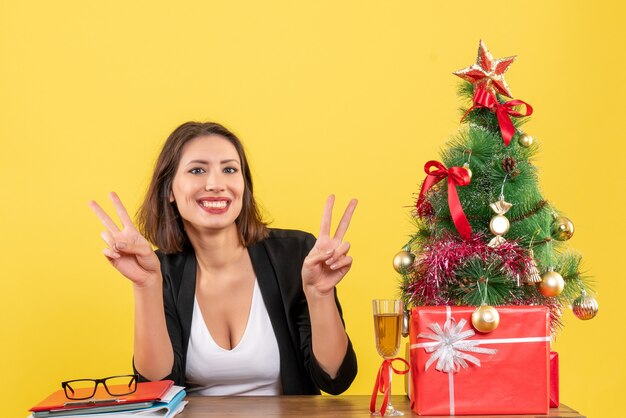 Estado de ánimo de año nuevo con hermosa dama de negocios haciendo gesto de victoria con los dedos y sentado en una mesa en la oficina