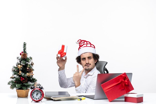 Estado de ánimo de año nuevo con joven empresario con sombrero de santa claus sentado en la oficina y sosteniendo su regalo mostrando dos sobre fondo blanco.