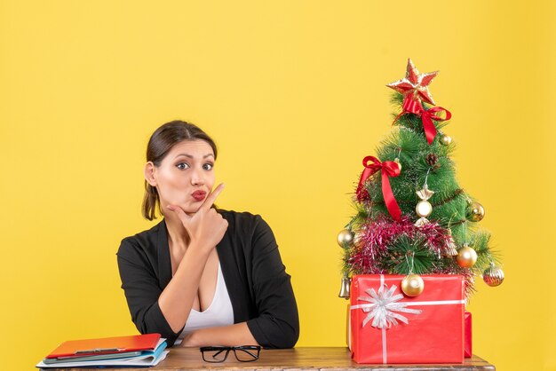 Estado de ánimo de año nuevo con joven sorprendida en traje con árbol de Navidad decorado en la oficina en amarillo