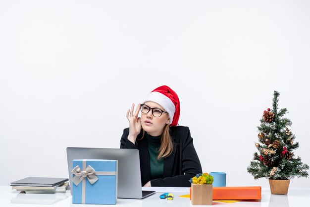 Estado de ánimo de año nuevo con mujer atractiva confundida con un sombrero de santa claus sentado en una mesa con un árbol de Navidad y un regalo en la oficina