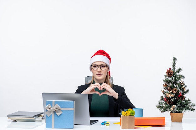 Estado de ánimo de año nuevo con una mujer atractiva romántica con un sombrero de santa claus sentado en una mesa con un árbol de Navidad y un regalo sobre fondo blanco