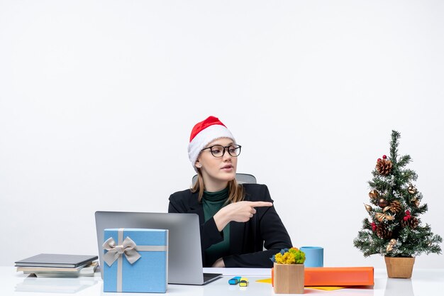 Estado de ánimo de año nuevo con una mujer joven y atractiva con un sombrero de santa claus sentado en una mesa con un árbol de Navidad y un regalo apuntando algo en la oficina