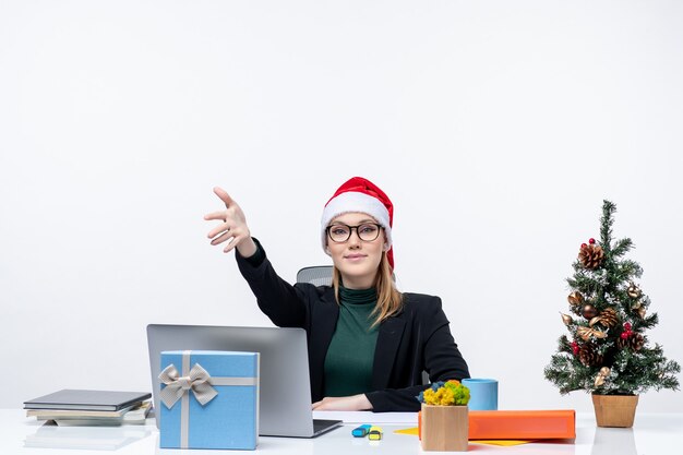 Estado de ánimo de año nuevo con una mujer joven y atractiva con un sombrero de santa claus sentado en una mesa con un árbol de Navidad y un regalo dando la bienvenida a alguien en la oficina