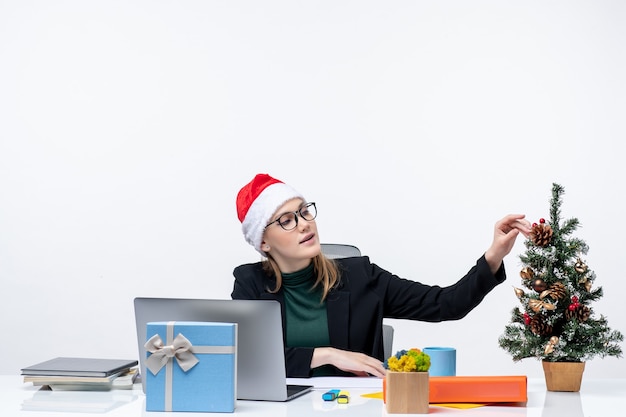 Estado de ánimo de año nuevo con una mujer joven y atractiva con un sombrero de santa claus sentado en una mesa con un árbol de Navidad y un regalo sobre fondo blanco.