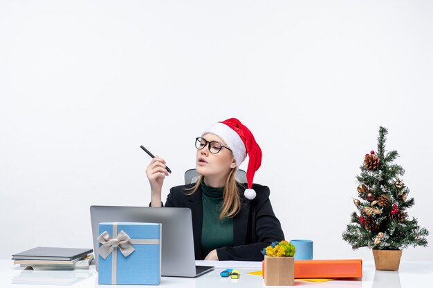 Estado de ánimo de año nuevo con mujer rubia concentrada con un sombrero de santa claus sentado en una mesa con un árbol de Navidad y un regalo sobre fondo blanco