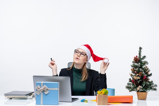 Estado de ánimo de año nuevo con mujer rubia de ensueño con un sombrero de santa claus sentado en una mesa con un árbol de Navidad y un regalo sobre fondo blanco