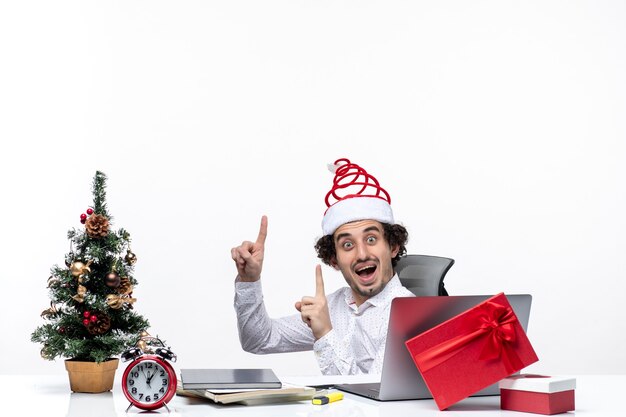 Estado de ánimo de año nuevo con sorprendido sonriente joven empresario emocionado sentado en la oficina y apuntando arriba sobre fondo blanco.