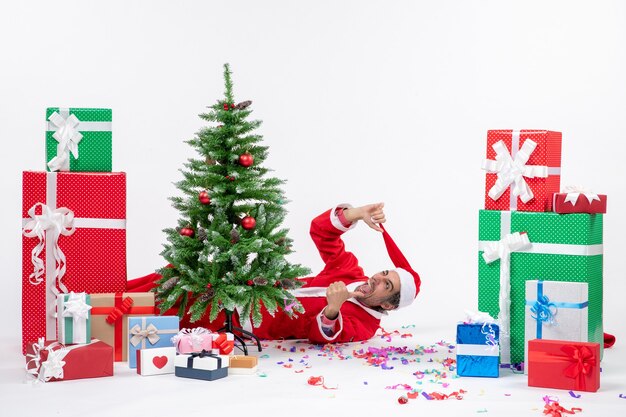 Estado de ánimo de celebración festiva con joven Papá Noel loco acostado detrás del árbol de Navidad cerca de regalos sobre fondo blanco.