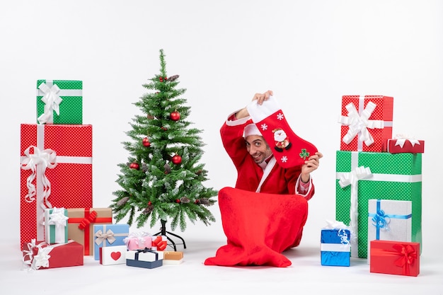 Estado de ánimo festivo con divertido santa claus sorprendido positivo sentado en el suelo y mostrando el calcetín de Navidad cerca de regalos y árbol de Navidad decorado sobre fondo blanco.