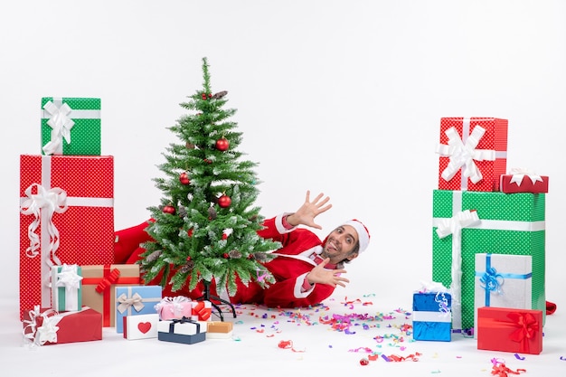 Estado de ánimo festivo con el joven Papá Noel acostado detrás del árbol de Navidad cerca de regalos sobre fondo blanco.