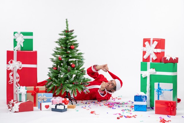 Foto gratuita estado de ánimo festivo con joven santa claus divertido acostado detrás del árbol de navidad cerca de regalos sobre fondo blanco foto de stock