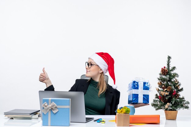 Estado de ánimo festivo con una mujer joven sorprendida con sombrero de santa claus y gafas sentado en una mesa que muestra el regalo de Navidad apuntando algo en el lado derecho haciendo un gesto de ok