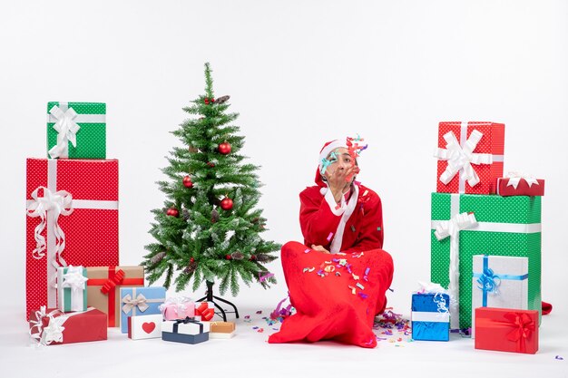 Estado de ánimo festivo con Papá Noel positivo divertido sentado en el suelo y jugando con adornos navideños cerca de regalos y árbol de Navidad decorado sobre fondo blanco.