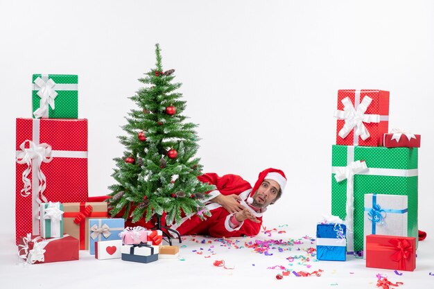 Estado de ánimo festivo con santa claus acostado detrás del árbol de navidad cerca de regalos en diferentes colores sobre fondo blanco.