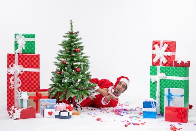 Estado de ánimo festivo con santa claus acostado detrás del árbol de navidad cerca de regalos en diferentes colores sobre fondo blanco.