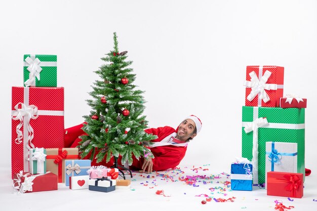 Estado de ánimo festivo con santa claus acostado detrás del árbol de navidad cerca de regalos sobre fondo blanco.