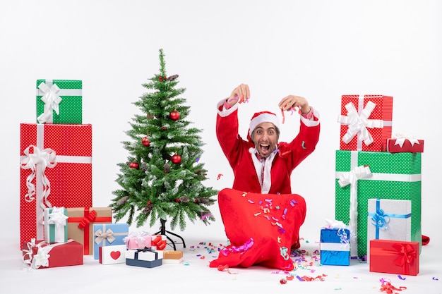 Estado de ánimo festivo con santa claus feliz positivo divertido sentado en el suelo y jugando con adornos navideños cerca de regalos y árbol de Navidad decorado sobre fondo blanco