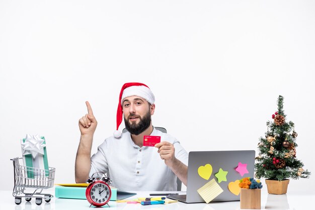 Estado de ánimo de Navidad con un adulto joven con sombrero de santa claus y sosteniendo una tarjeta bancaria y apuntando hacia arriba en la oficina