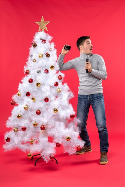 Estado de ánimo de Navidad con un chico seguro de pie cerca del árbol de Navidad decorado y sosteniendo el micrófono tomando una foto y mirando algo