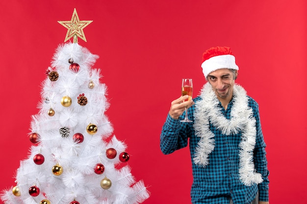 Estado de ánimo de Navidad con confianza emocional decidido joven con sombrero de santa claus y levantando una copa de vino cerca del árbol de Navidad