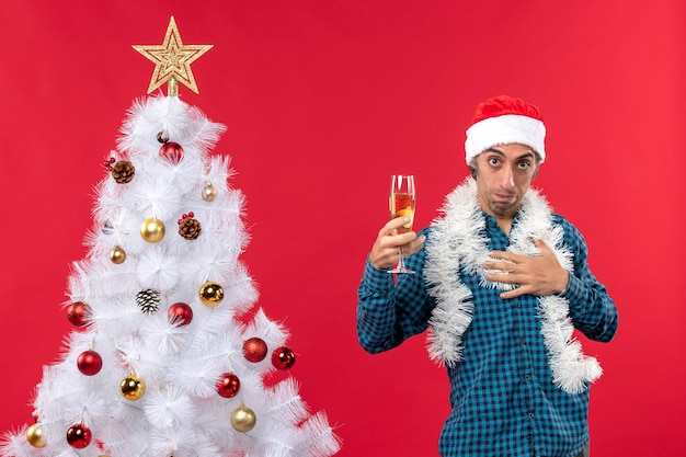 Estado de ánimo de Navidad con confianza joven con sombrero de santa claus en una camisa azul a rayas sosteniendo una copa de vino cerca del árbol de Navidad