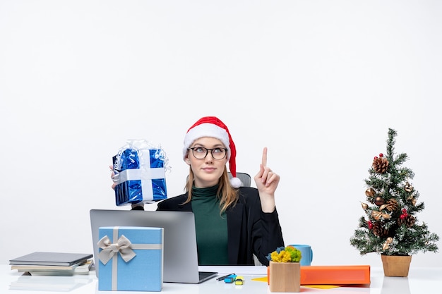 Estado de ánimo de Navidad con curiosa mujer joven con sombrero de santa claus y gafas sentado en una mesa sosteniendo su regalo sorprendentemente sobre fondo blanco.