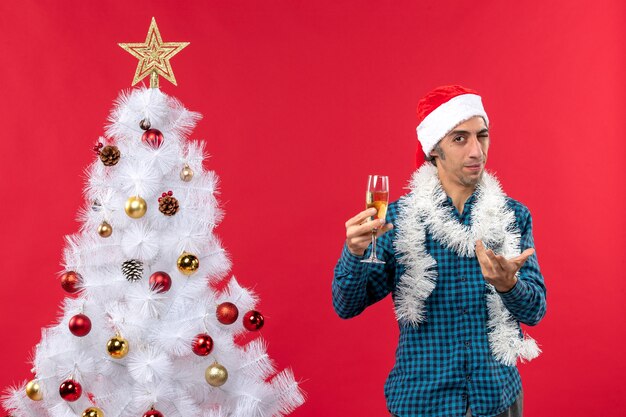 Estado de ánimo de Navidad con curioso joven con sombrero de santa claus en una camisa azul a rayas levantando una copa de vino cerca del árbol de Navidad