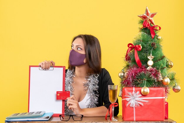 Estado de ánimo de Navidad con dama encantadora en traje con máscara médica con documento y tarjeta bancaria en la oficina en amarillo aislado