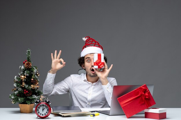 Estado de ánimo de Navidad con divertida persona de negocios con sombrero de santa claus levantando su regalo a la cara y haciendo gesto de anteojos sobre fondo blanco.