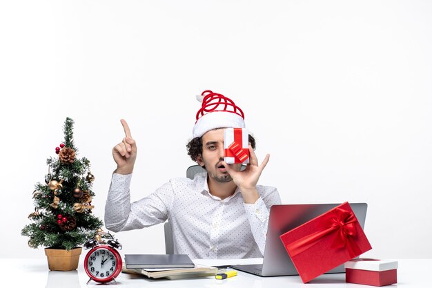 Estado de ánimo de Navidad con divertida persona de negocios con sombrero de santa claus levantando su regalo a su cara y apuntando arriba sobre fondo blanco.