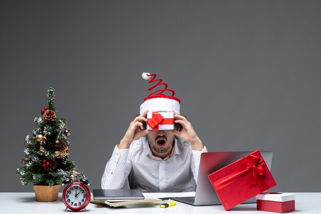 Estado de ánimo de Navidad con divertida persona de negocios con sombrero de santa claus sosteniendo su regalo delante de su cara sobre fondo oscuro