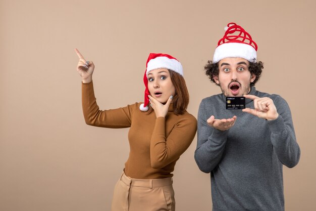 Estado de ánimo de Navidad con emocionado pareja fresca satisfecha vistiendo sombreros rojos de santa claus chico mostrando tarjeta bancaria, mujer apuntando arriba