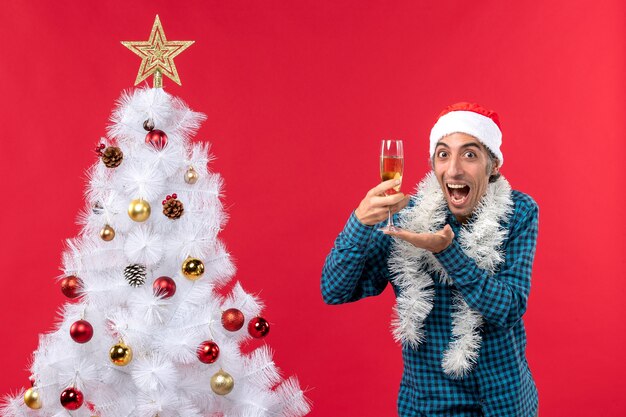 Estado de ánimo de Navidad con emocional feliz gracioso joven con sombrero de santa claus en una camisa azul a rayas sosteniendo una copa de vino cerca del árbol de Navidad
