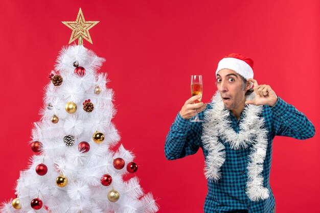 Estado de ánimo de Navidad con emocional joven con sombrero de santa claus en una camisa azul a rayas escuchando atentamente sosteniendo una copa de vino cerca del árbol de Navidad