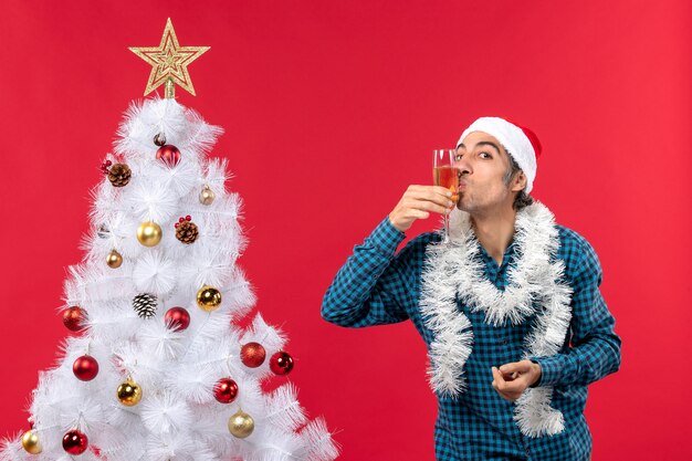 Estado de ánimo de Navidad con emocional joven con sombrero de santa claus en una camisa azul a rayas sosteniendo y besando copa con vino cerca del árbol de Navidad