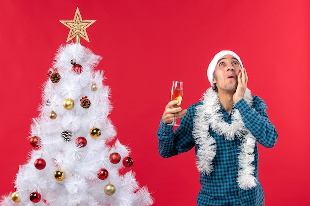 Estado de ánimo de Navidad con emocional joven con sombrero de santa claus en una camisa azul a rayas sosteniendo una copa de vino mirando hacia abajo cerca del árbol de Navidad