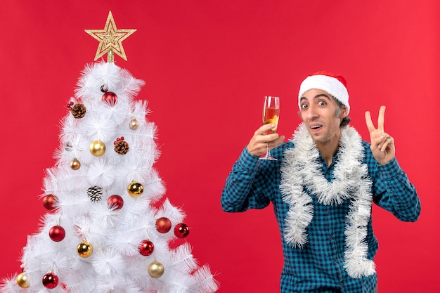 Estado de ánimo de Navidad con emocional joven con sombrero de santa claus en una camisa azul a rayas sosteniendo una copa de vino y mostrando dos cerca del árbol de Navidad