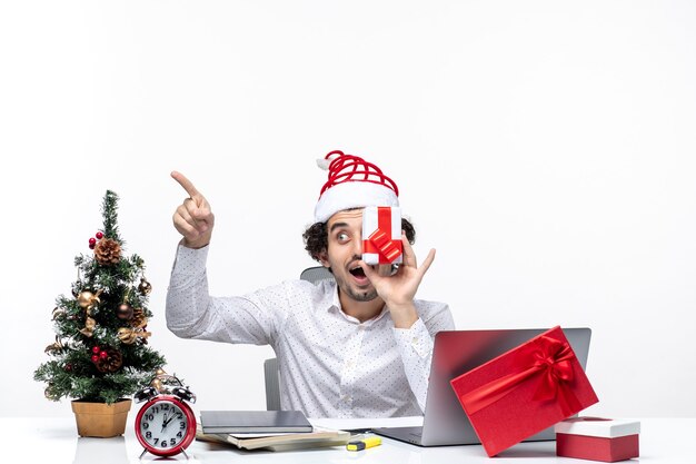 Estado de ánimo de Navidad con empresario sorprendido con sombrero de santa claus levantando su regalo a la cara y apuntando algo sobre fondo blanco.