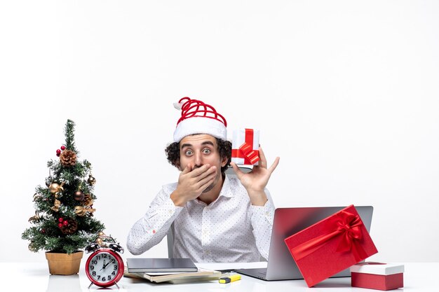 Estado de ánimo de Navidad con empresario sorprendido con sombrero de santa claus levantando su regalo y cerrando la boca sobre fondo blanco.