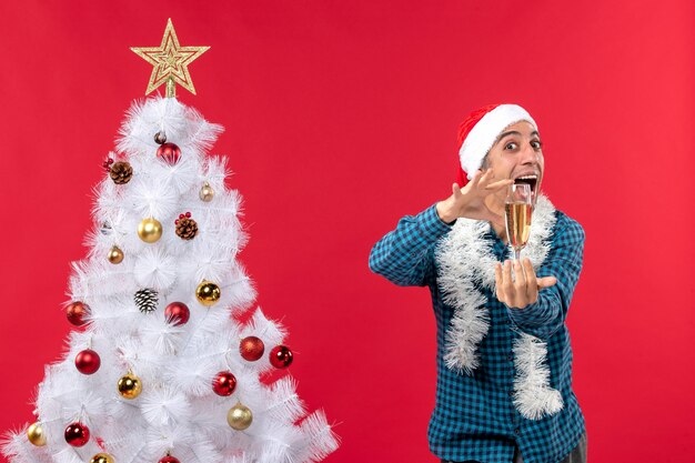 Estado de ánimo de Navidad con feliz joven con sombrero de santa claus en una camisa azul a rayas levantando una copa de vino cerca del árbol de Navidad