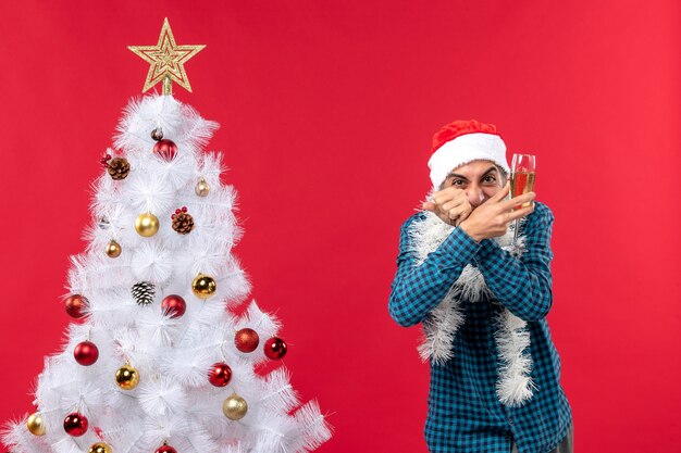 Estado de ánimo de Navidad con feliz loco emocional joven con sombrero de santa claus en una camisa azul a rayas levantando una copa de vino cerca del árbol de Navidad