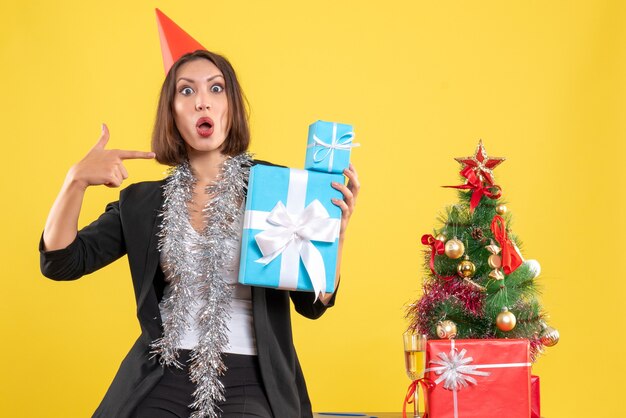 Estado de ánimo de Navidad con hermosa dama emocional con sombrero de Navidad señalando regalos en la oficina en amarillo