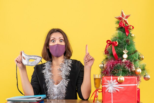 Estado de ánimo de Navidad con hermosa dama pensativa en traje con su máscara médica apuntando hacia arriba en la oficina