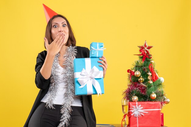 Estado de ánimo de Navidad con hermosa dama sorprendida emocional con sombrero de Navidad con regalos en la oficina en amarillo