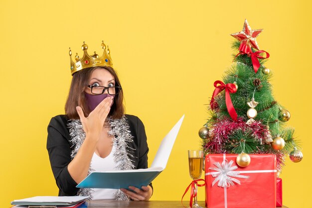 Estado de ánimo de Navidad con una hermosa dama sorprendida en traje con corona con su máscara médica sosteniendo documentos en la oficina