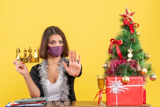 Estado de ánimo de Navidad con hermosa dama en traje con corona de sujeción con su máscara médica haciendo gesto de parada en la oficina en amarillo