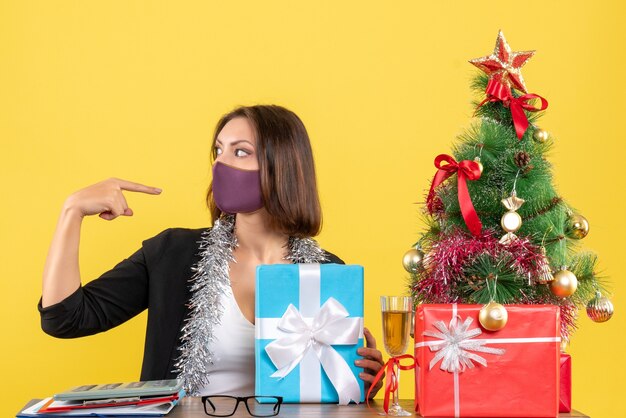 Estado de ánimo de Navidad con hermosa dama en traje con máscara médica y sosteniendo un regalo apuntando a sí misma en la oficina en amarillo