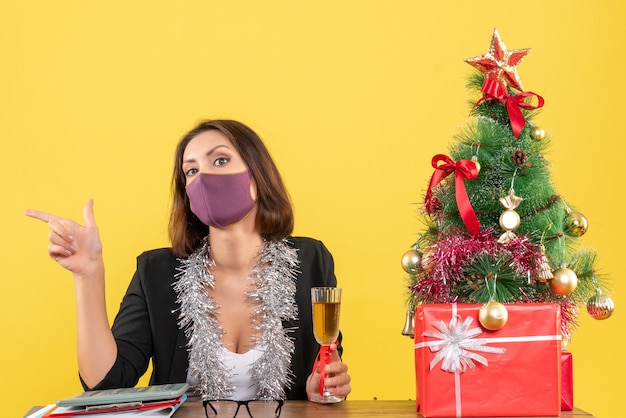 Estado de ánimo de Navidad con hermosa dama en traje con máscara médica y sosteniendo vino en la oficina en amarillo