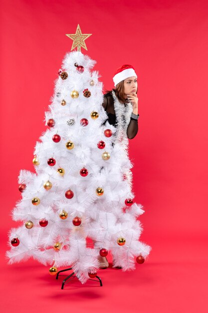 Estado de ánimo de Navidad con una hermosa joven sorprendida en un vestido negro con sombrero de santa claus escondido detrás del árbol de año nuevo